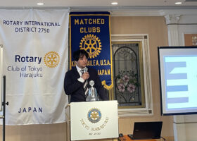 東京原宿ロータリークラブの演台でマイクを持って講演する様子